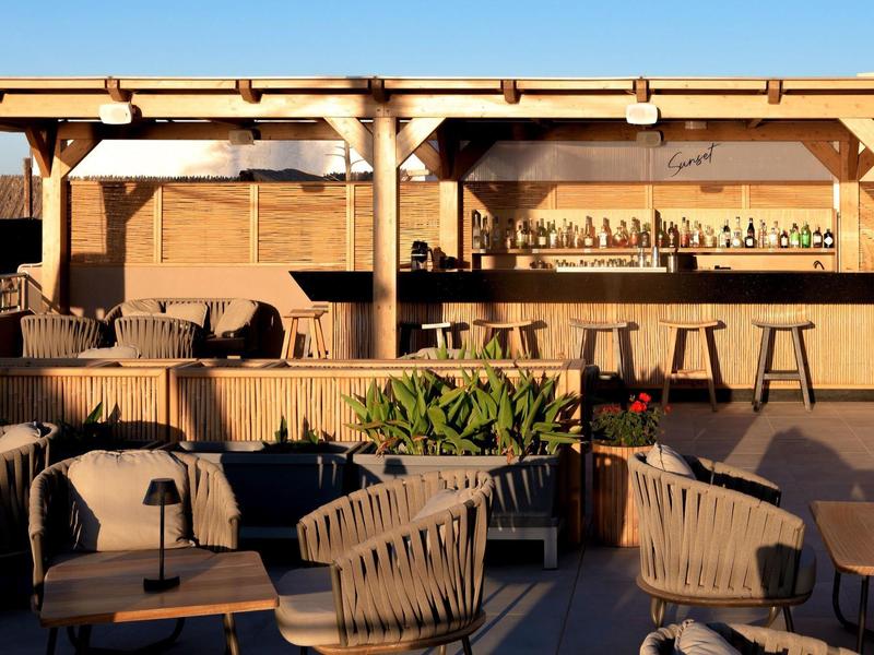 Zewnętrzny bar z drewnianymi meblami, stołkami i roślinami w ciepłym świetle słonecznym.