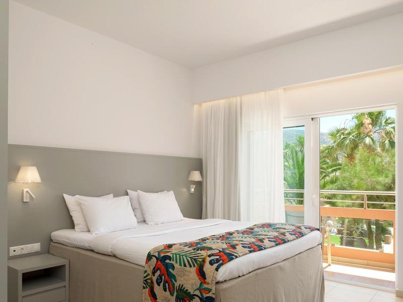 Jasny pokój hotelowy z dużym łóżkiem, lampkami nocnymi i balkonem z tropikalną roślinnością.