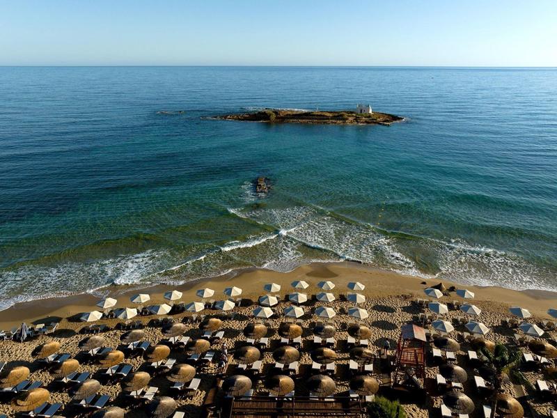 Plaża z rzędami parasoli i leżaków nad spokojnym niebieskim morzem i małą skalistą wyspą.