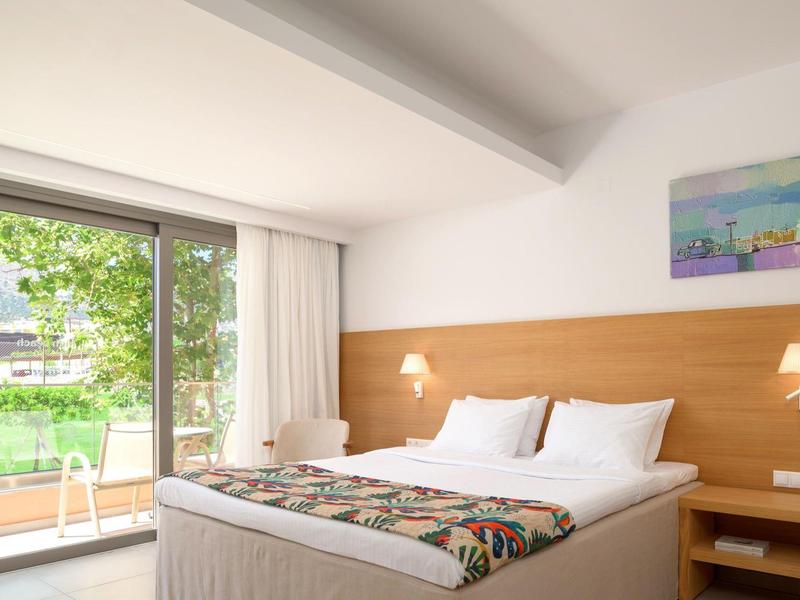 Jasny pokój hotelowy z dużym łóżkiem, drewnianym zagłówkiem i widokiem na ogród przez szklane drzwi.