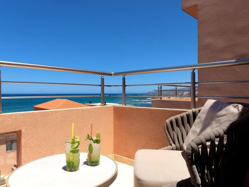 Przytulny balkon z poduszkowanym fotelem, małym stolikiem, napojami, widokiem na morze pod bezchmurnym niebem.