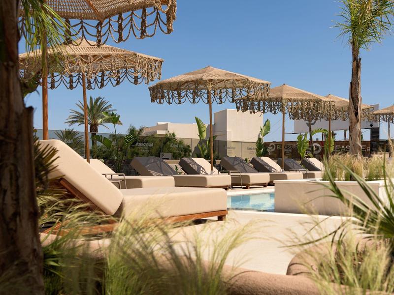 Leżaki i słomiane parasole przy czystym, niebieskim basenie z palmami.