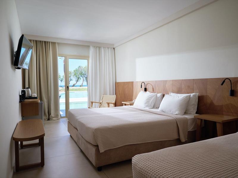 Hell eingerichtetes Hotelzimmer mit Doppelbett, Einzelbett, Fernseher und Blick auf Balkon mit Meer.