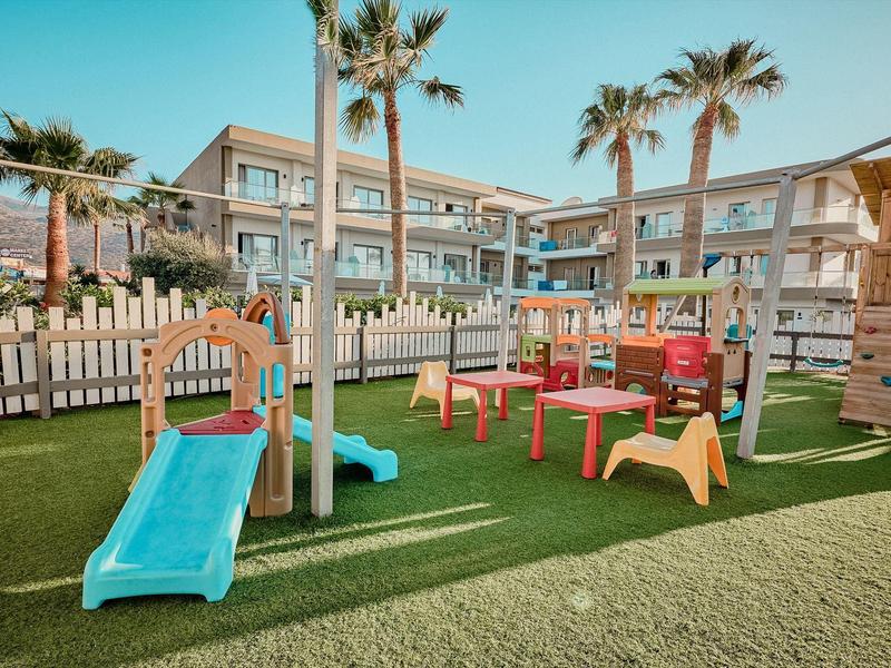 Ein bunter Kinderspielplatz mit Rutsche, Tischen, Stühlen und Palmen vor einem Gebäude.