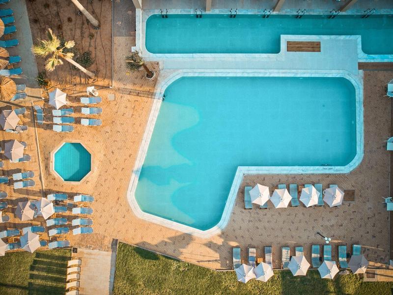 Vogelperspektive auf blauen Pool mit Liegestühlen, Sonnenschirmen und Sandboden daneben.