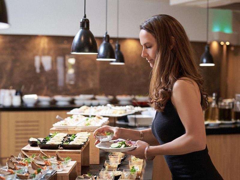 Eine Frau in schwarzem Kleid nimmt Speisen von einem langen Buffet mit vielen kleinen Gläsern und Schüsseln.