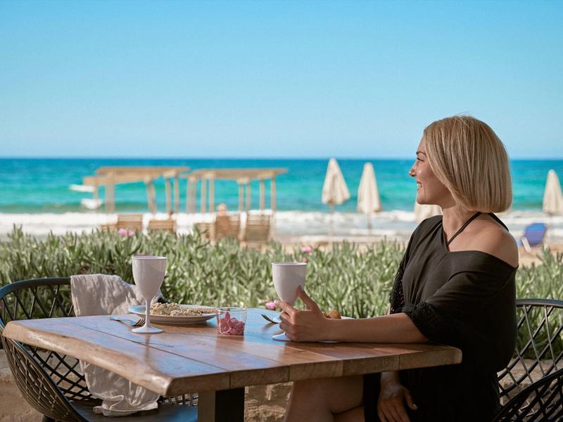 Frau sitzt entspannt am Tisch mit Laptop und Getränk, Meer und Strand mit Sonnenschirmen im Hintergrund.