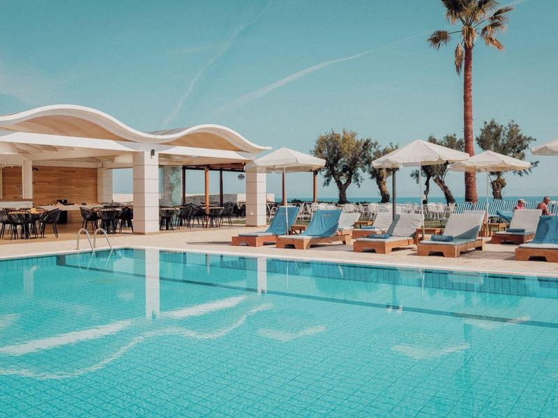 Ein Pool mit kristallklarem Wasser, Liegestühlen und Palmen am sonnigen Meerufer.