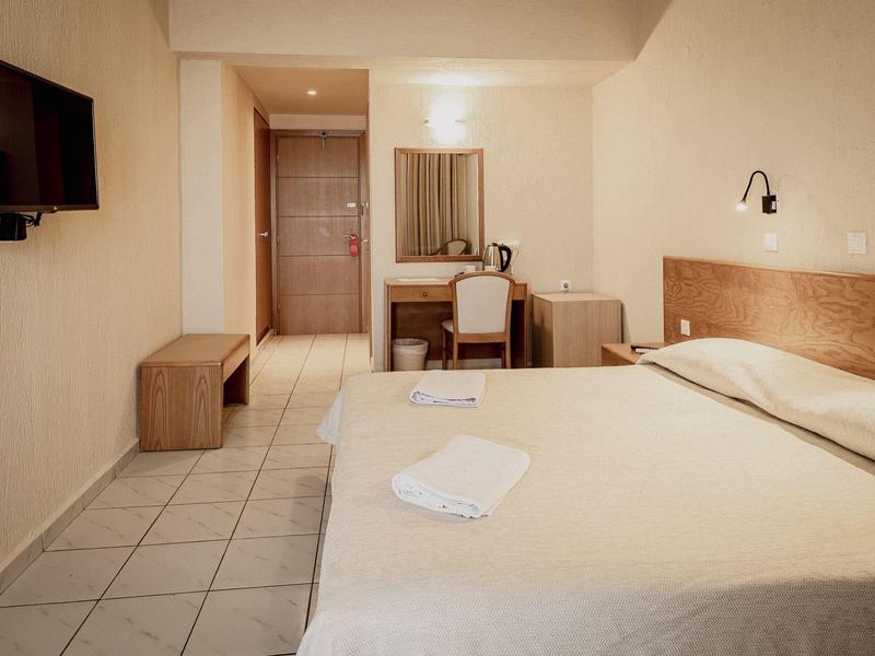 Einfaches Hotelzimmer mit Doppelbett, weißer Bettwäsche, Fernseher und Schreibtisch vor heller Wand.