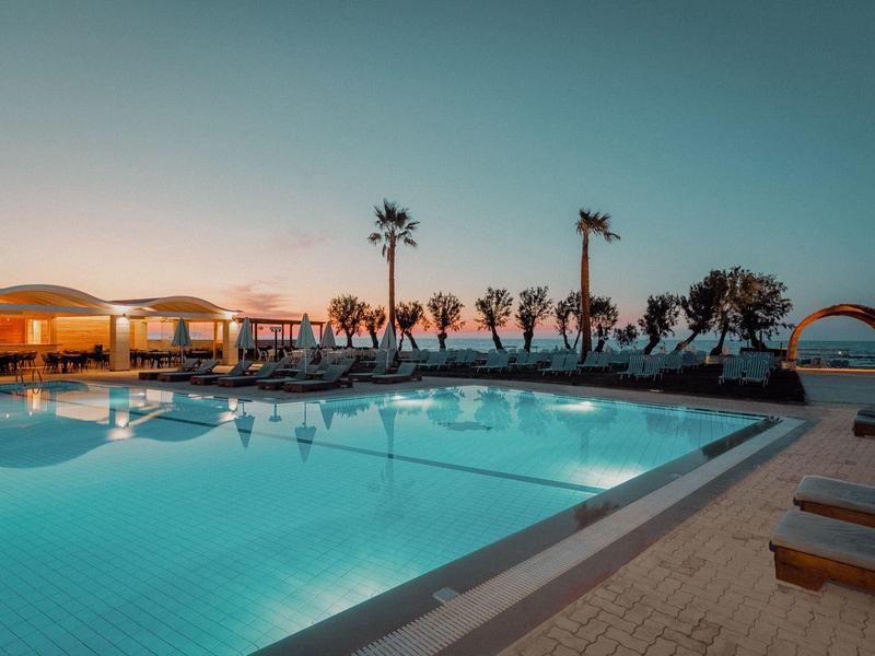 Großer beleuchteter Pool bei Sonnenuntergang mit Palmen und Liegestühlen am Meer.