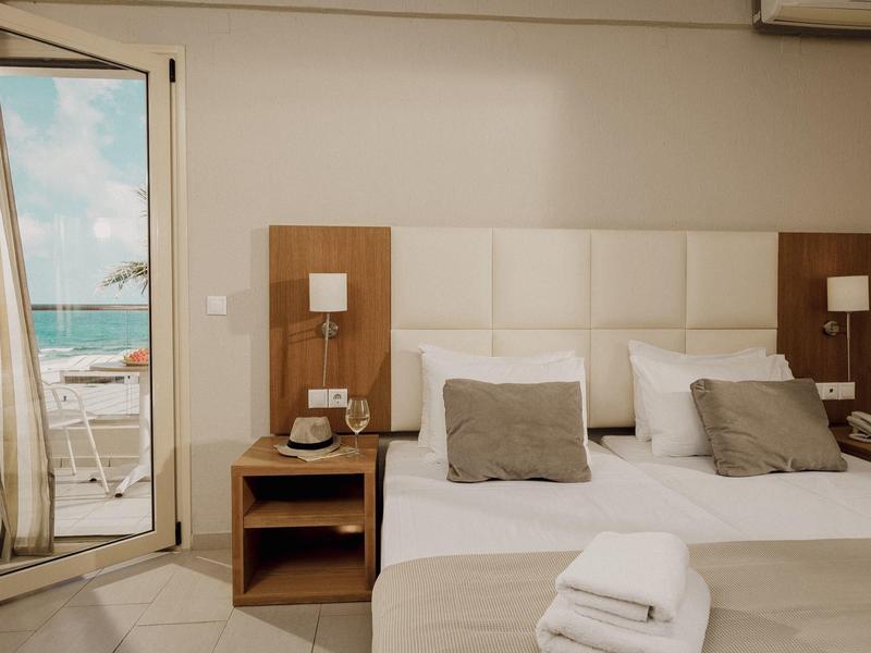 Hell eingerichtetes Schlafzimmer mit Doppelbett, zwei Nachttischen, Lampen und Blick auf Meer durch offene Tür.