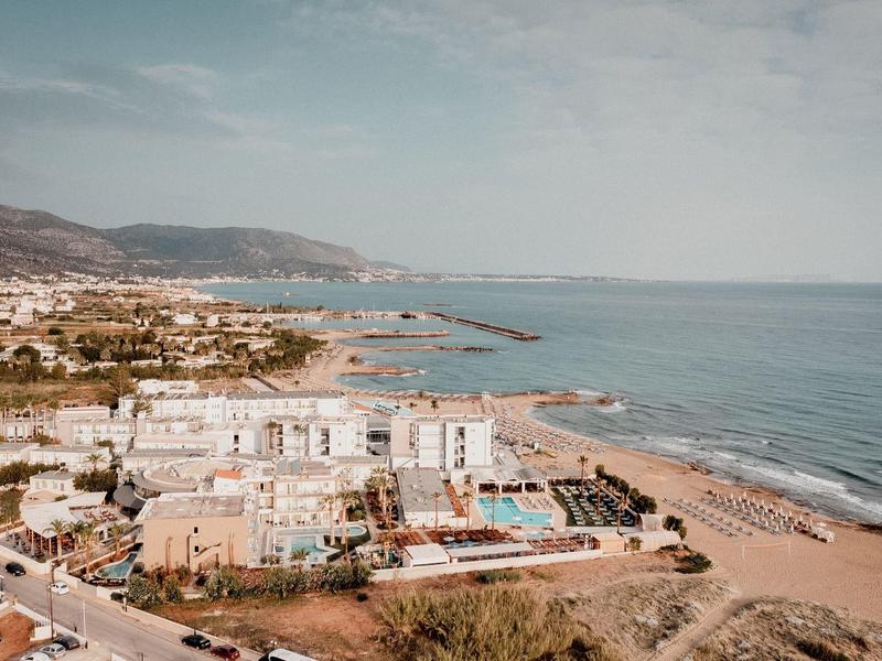 Küstenstadt mit Sandstrand, Hotels, Palmen und Bergen im Hintergrund unter blauem Himmel.