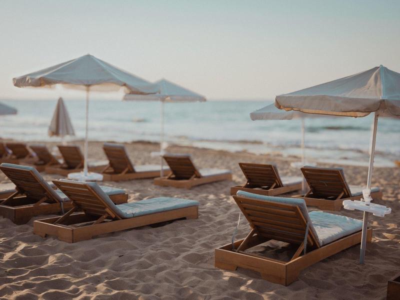 Leere Holzbettliegen mit weißen Auflagen und Sonnenschirmen am sandigen Strand bei ruhigem Meer.