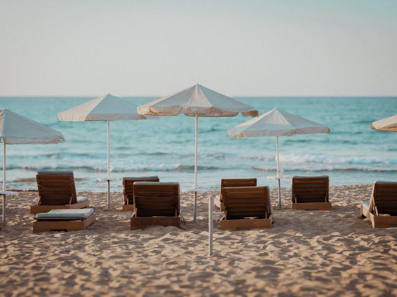 Strand mit braunen Liegestühlen und hellen Sonnenschirmen vor ruhigem, blauem Meer bei klarem Himmel.