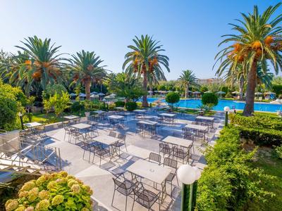 Area pranzo all'aperto con tavoli e sedie accanto a una piscina circondata da palme.