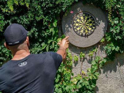 Mann spielt Darts vor einer von Grün bewachsenen Wand.