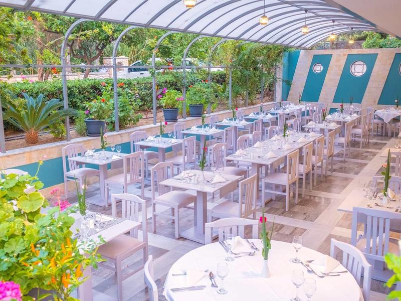 Jasna zadaszona przestrzeń na zewnątrz z białymi stołami i krzesłami, duży basen i bujny widok na ogród.