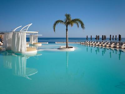 Large pool with palm island, sun loungers, and sea in the background under clear sky.