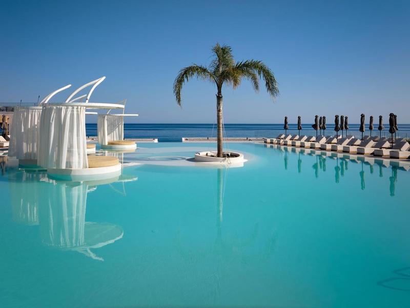 Groot zwembad met palmeneiland en ligstoelen onder blauwe lucht aan zee