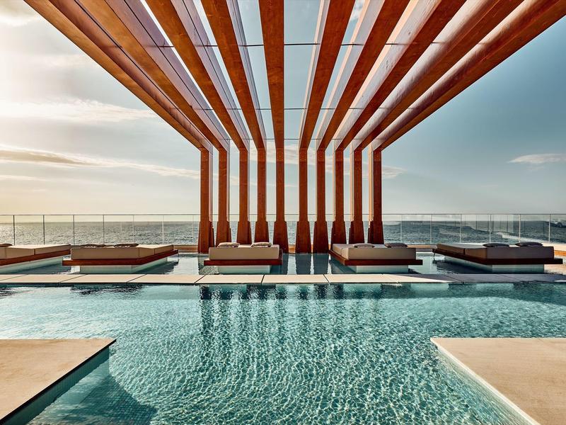 Moderne zwembadruimte met houten balken en ligstoelen aan zee bij zonsondergang.