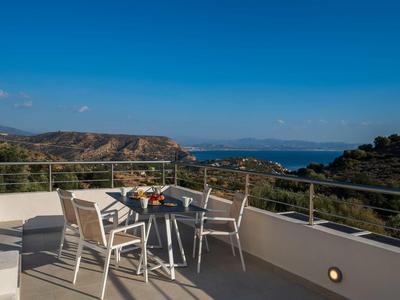 Terrasse avec table et chaises donnant sur des collines et la mer sous un ciel bleu.