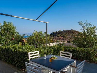 Terrasse avec table et chaises blanches, plantes vertes et collines en arrière-plan sous un ciel clair.