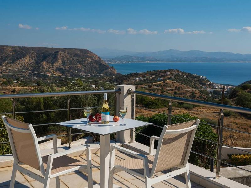 Terrasse avec table et chaises offrant une vue sur les collines et la mer sous un ciel bleu.