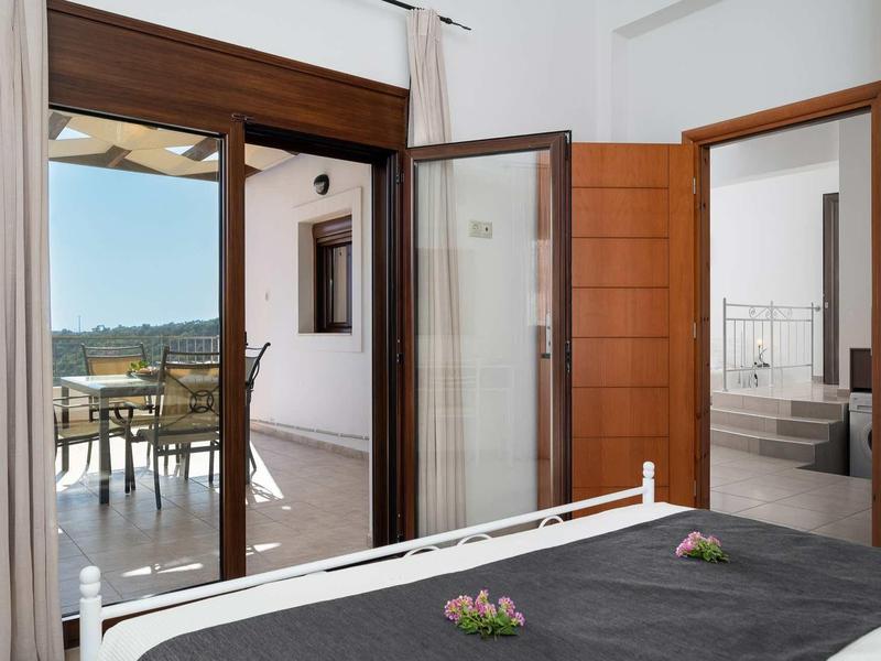 Vue depuis une chambre avec un lit double sur une terrasse avec table et chaises par temps ensoleillé.