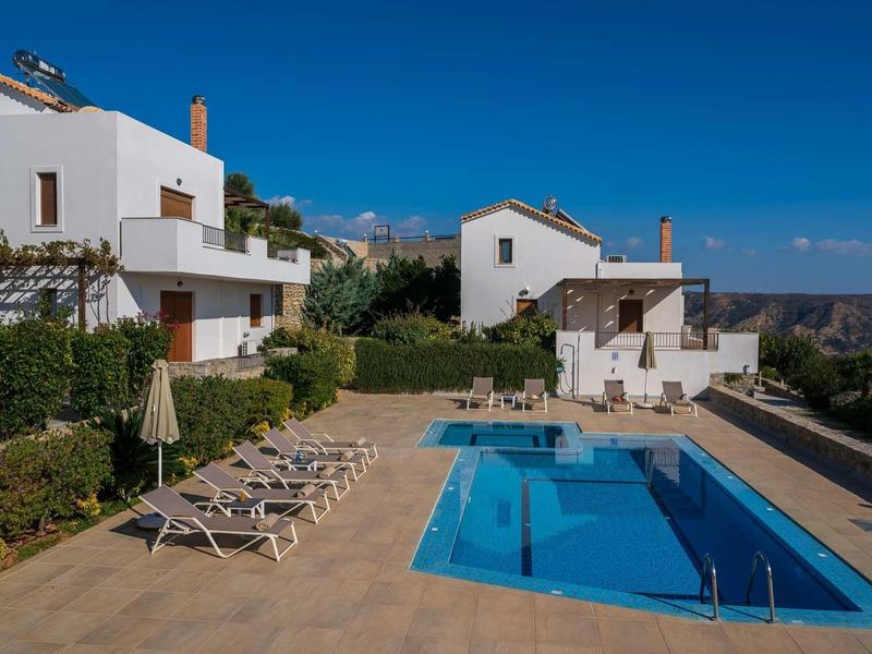 Deux maisons de vacances blanches avec terrasse, piscine et plusieurs chaises longues sous un ciel bleu clair.