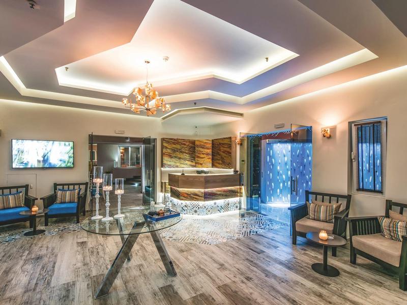 Spacious hotel lobby with modern chandelier, wooden furniture, and backlit reception desk.