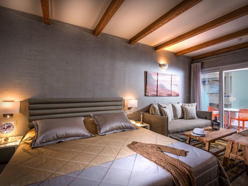 Cozy hotel room with a double bed, sofa, exposed wooden beams, and a balcony with outdoor seating.