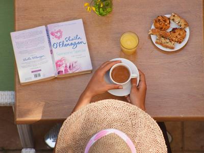 Osoba w kapeluszu słomkowym siedzi przy drewnianym stole z książką, napojami i ciastkami.