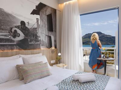 Jasny pokój hotelowy z białym łóżkiem i balkonem z widokiem na góry i wodę.