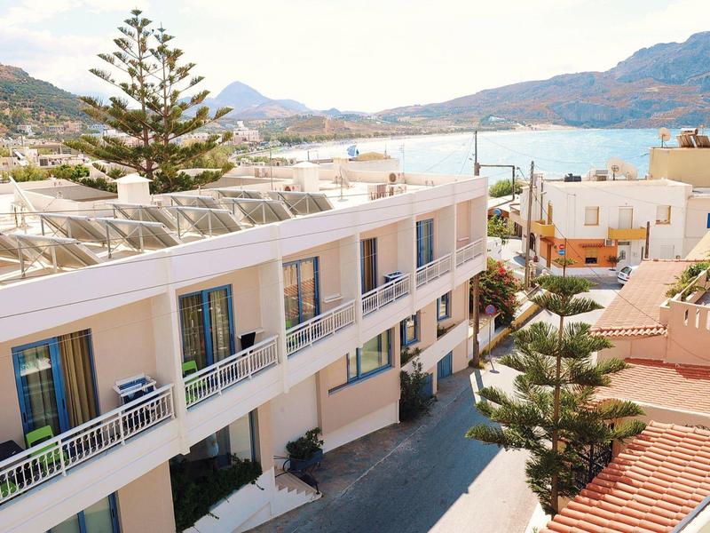 Widok na biały hotel z balkonami nad zatoką otoczoną zielonymi wzgórzami.