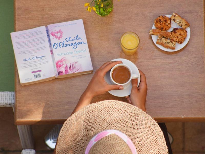 Osoba z kapeluszem słomkowym pije kawę przy stole z książkami, szklanką i ciastkami.