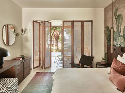 Jasny pokój hotelowy z łóżkiem, drewnianymi meblami i widokiem na taras z palmami.