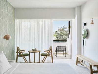 Helles Schlafzimmer mit grünem Akzent, Holzstühlen, kleinem Tisch und Balkon mit Außenstuhl.