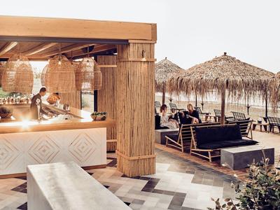 Strandbar mit Holzmöbeln, Strohdächern und Sitzgelegenheiten, Meer im Hintergrund bei Sonnenuntergang.