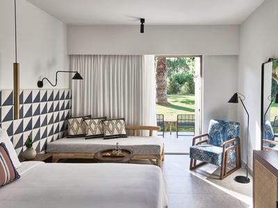 Moderner Raum mit weißem Bett, geometrischer Wandkunst, Holzbank, blauem Sessel und Gartenausblick.