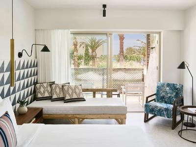 Helles Schlafzimmer mit weißer Wand, gepolstertem Bett, Holzbank, blauem Sessel und großem Fenster.