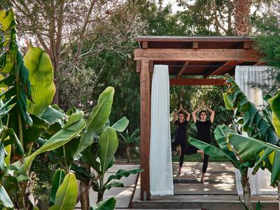 Zwei Personen im Yoga-Pose vor Holzbogen, umgeben von großen grünen Blättern und Bäumen.