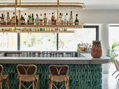 Moderne Bar mit grüner Fliesenfront, weißen Arbeitsflächen und Rattanstühlen, beleuchtet von goldfarbenen Hängelampen.