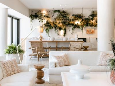 Helles Wohnzimmer mit weißen Sofas, Holzmöbeln, großen Fenstern und grünen Pflanzen an der Wand.