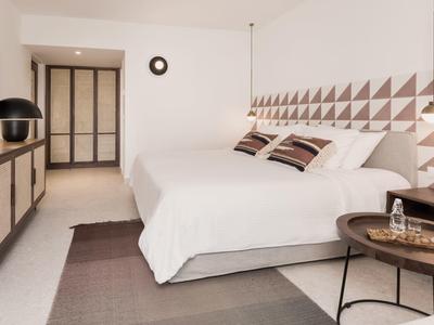 Nowoczesny pokój hotelowy z dużym łóżkiem, geometrycznym wzorem na ścianie i stylowymi stolikami nocnymi.