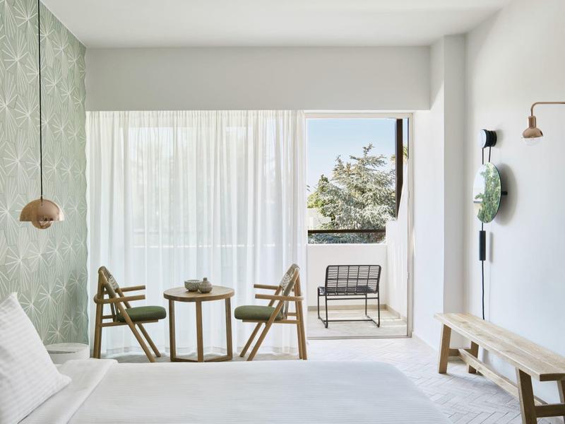 Jasny pokój hotelowy z podwójnym łóżkiem, dwoma drewnianymi krzesłami, małym stolikiem i balkonem z widokiem na morze i drzewa.