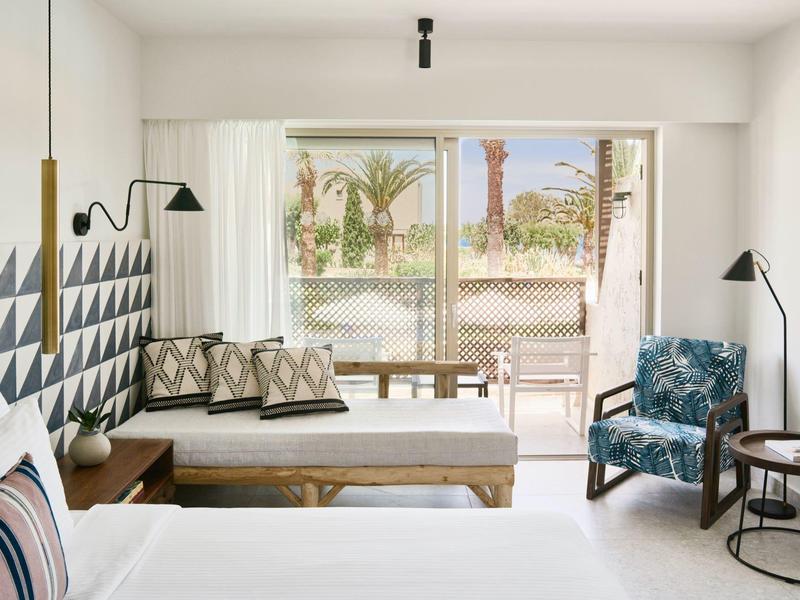 Jasny pokój hotelowy z nowoczesnym wystrojem, łóżkiem, miejscem do siedzenia i balkonem z widokiem na palmy.