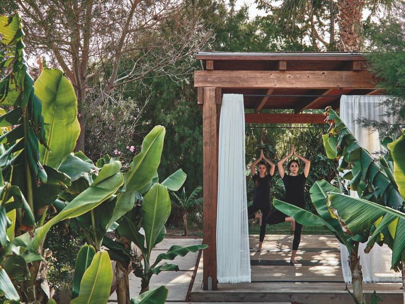 Dwie kobiety ćwiczą jogę na drewnianej platformie otoczonej tropikalnymi roślinami.