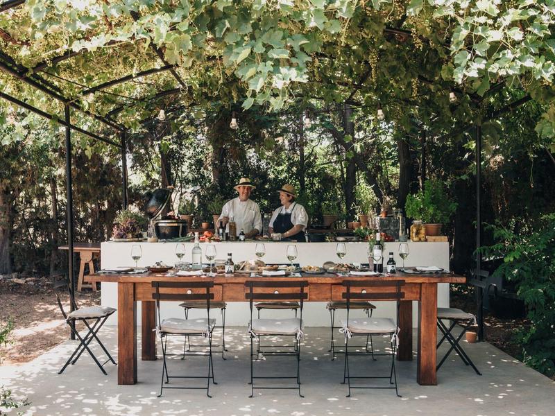 Przestrzeń do jedzenia na zewnątrz pod liściastym daszkiem z stołem, krzesłami i dwoma osobami w tle.