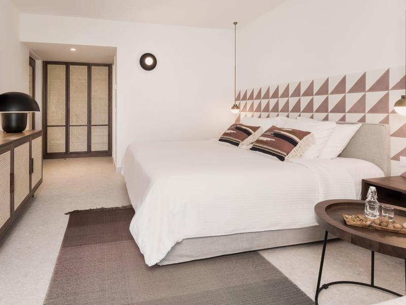 Nowoczesny pokój hotelowy z dużym łóżkiem, geometrycznym wzorem na ścianie i stylowymi stolikami nocnymi.