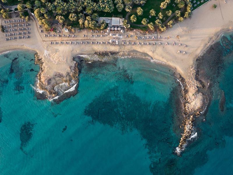Luftaufnahme eines Strandes mit klar türkisfarbenem Wasser und schattigen Bereichen im Sand